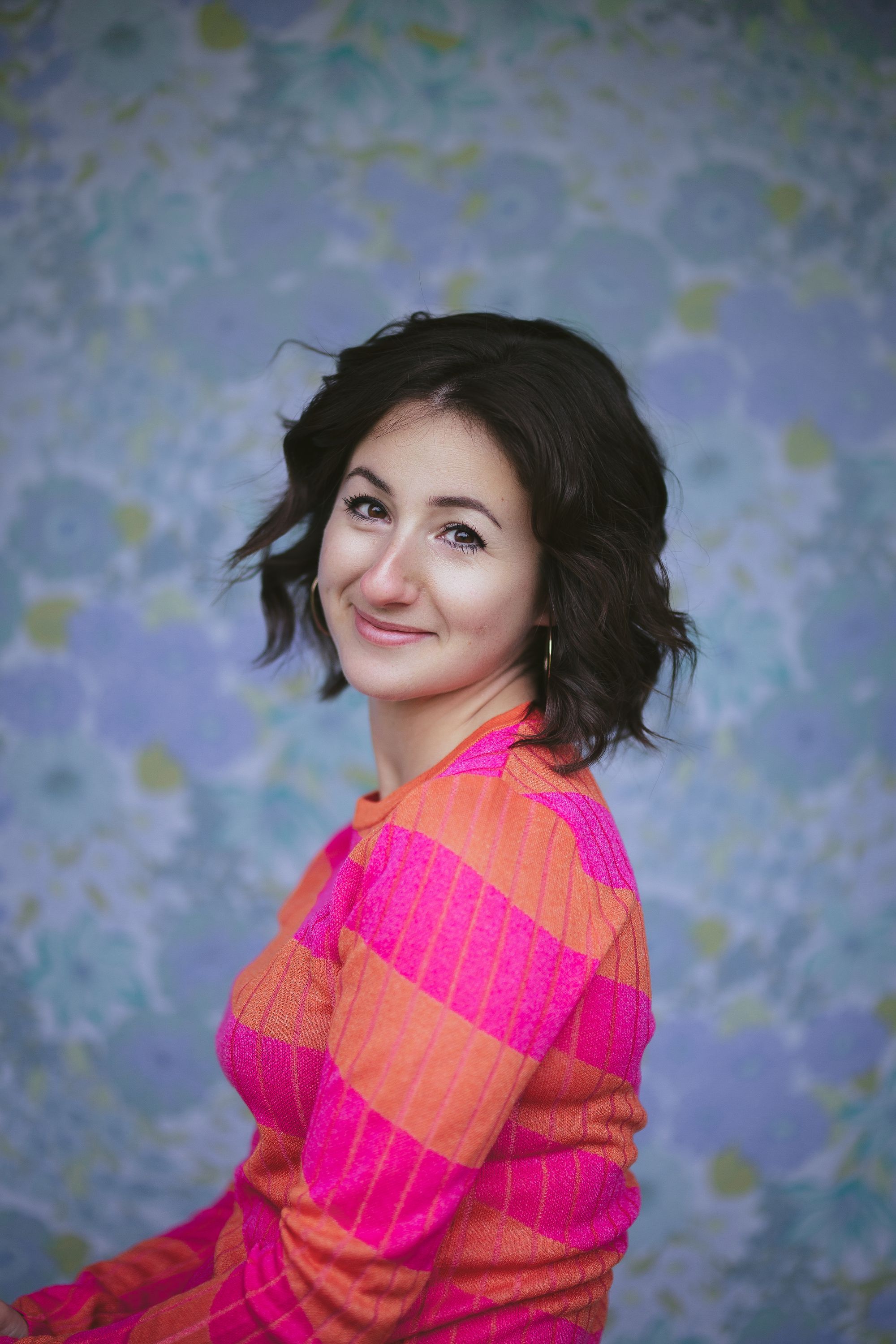 A portrait photograph of me sitting, smiling at the photographer, wearing a pink and orange striped top